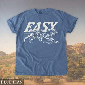Op de afbeelding: Blauw spijkerbroekkleurig T-shirt met het woord "EASY" in witte letters boven een witte tijgerafbeelding. De tijger wordt lopend afgebeeld met zichtbare strepen. De mouwen van het shirt zijn opgerold.