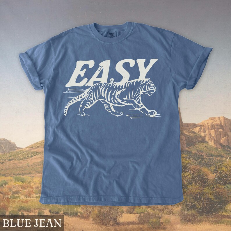 Op de afbeelding: Blauw spijkerbroekkleurig T-shirt met het woord "EASY" in witte letters boven een witte tijgerafbeelding. De tijger wordt lopend afgebeeld met zichtbare strepen. De mouwen van het shirt zijn opgerold.