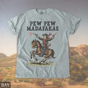 Puede incluir: Camiseta azul claro con un dise&ntilde;o de vaquero. El vaquero, montado a caballo, dispara dos pistolas. El texto "PEW PEW MADAFAKAS" est&aacute; impreso encima. La camiseta tiene mangas enrolladas y un corte relajado.