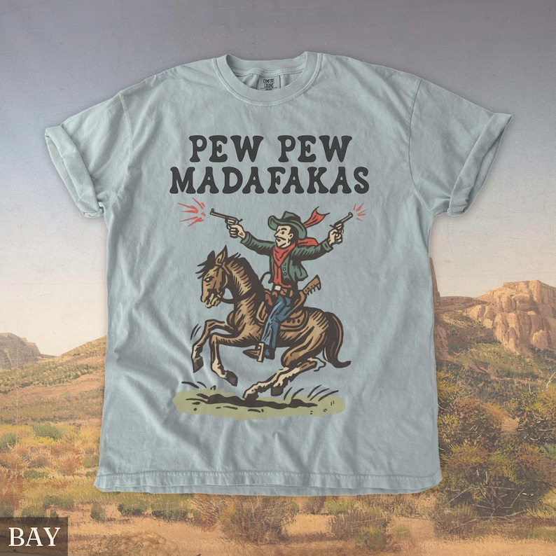 Puede incluir: Camiseta azul claro con un dise&ntilde;o de vaquero. El vaquero, montado a caballo, dispara dos pistolas. El texto "PEW PEW MADAFAKAS" est&aacute; impreso encima. La camiseta tiene mangas enrolladas y un corte relajado.