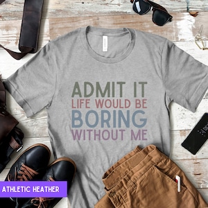 May include: Athletic heather gray t-shirt with the text "ADMIT IT LIFE WOULD BE BORING WITHOUT ME" in various colors. The shirt is displayed with brown pants, black shoes, and accessories on a wooden surface.