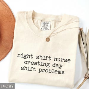 May include: Ivory-colored Comfort Colors t-shirt with the text "night shift nurse creating day shift problems" in black. A gold necklace with a pendant is draped across the shirt. A brown hat and dried plant are in the background.