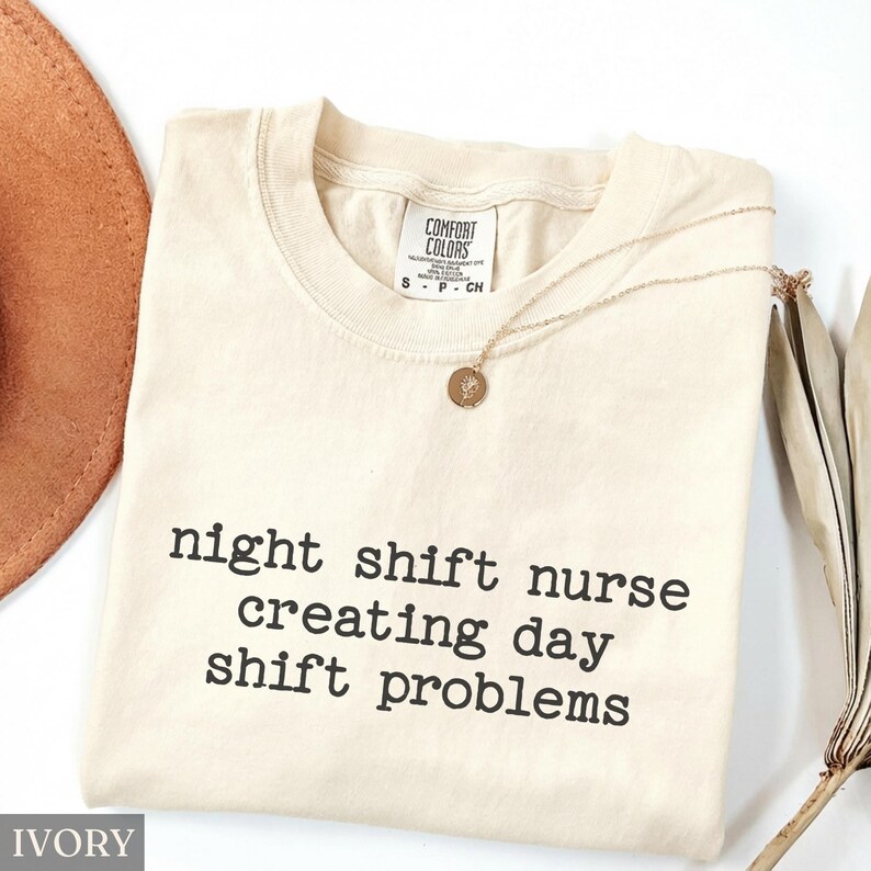 May include: Ivory-colored Comfort Colors t-shirt with the text "night shift nurse creating day shift problems" in black. A gold necklace with a pendant is draped across the shirt. A brown hat and dried plant are in the background.