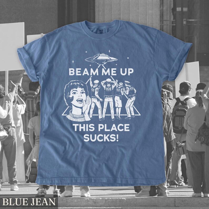 May include: Blue jean-coloured t-shirt with a graphic design. The design features a retro-style illustration of a woman and the text "BEAM ME UP THIS PLACE SUCKS!" along with an image of a UFO and figures.