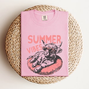 May include: Pink t-shirt with a graphic of a Highland cow wearing sunglasses and relaxing on a pool float. The text "Summer Vibes" is printed above the cow.