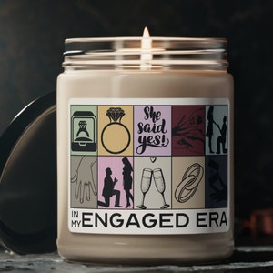 May include: A beige candle with a white label that says "In My Engaged Era" and features illustrations of rings, champagne flutes, and a couple getting engaged. The label also says "She said yes!"