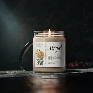 May include: A beige candle with a clear glass jar and a white label. The label features a chrysanthemum flower illustration and the text "Chrysanthemum, November Fourteenth, Abigail, As you blow out this candle, may all your wishes and dreams shine as brightly as its flame. Happy Birthday! Love, Mom".