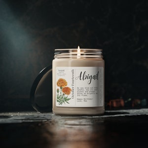 May include: A brown candle with a marigold flower design and the text "October Fourteenth" and "Abigail". The candle has a clear glass jar and a black lid. The text on the label reads "As you blow out this candle, may all your wishes and dreams shine as brightly as its flame. Happy Birthday! Love, Mom."