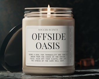 May include: A beige candle in a glass jar with a white label that reads "Soccer Scents Offside Oasis". The label also includes the text "Score a goal for tranquility and take a break from the chaos of the soccer field. Kick off your cleats, and let the stress of the game roll away."
