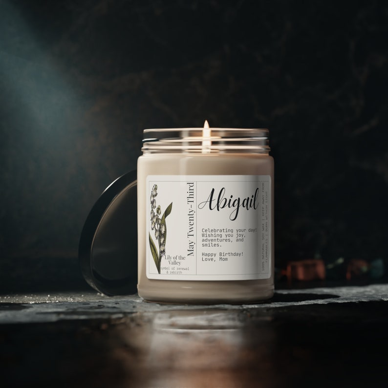 May include: A white candle with a brown label that says "Lily of the Valley" and "May Twenty-Third". The label also has a floral illustration and the text "Abigail", "Celebrating your day Wishing you joy, adventures, and smiles. Happy Birthday! Love, Mom".