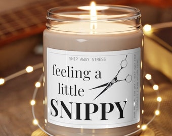 May include: A brown soy candle with a white label that reads "feeling a little SNIPPY" with a pair of scissors graphic. The candle is on a wooden surface with a string of white lights around it.