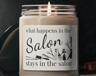 May include: A brown glass candle with a white label that reads "what happens in the Salon stays in the salon". The label also features a black and white illustration of salon tools, including scissors, combs, hairspray, and a bowl. The candle is lit and the flame is visible.