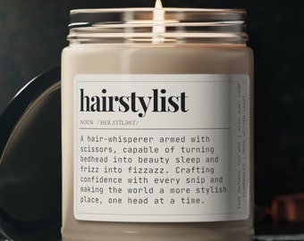 May include: A beige candle with a glass jar and a white label that reads "hairstylist" with the definition "NOUN /HER.STILIST/" and a description of a hairstylist. The candle is lit and the flame is visible.
