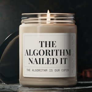 May include: A brown soy wax candle with a white label that reads "The Algorithm Nailed It" and "The Algorithm is our Cupid". The candle is lit and the flame is visible.