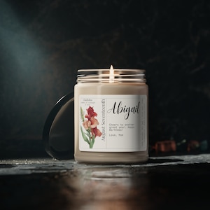 May include: A beige candle with a white label featuring a floral illustration and the text "Gladiolus Symbol of Strength & Love" and "August Seventeenth". The label also includes the name "Abigail" and the message "Cheers to another great year. Happy Birthday! Love, Mom".