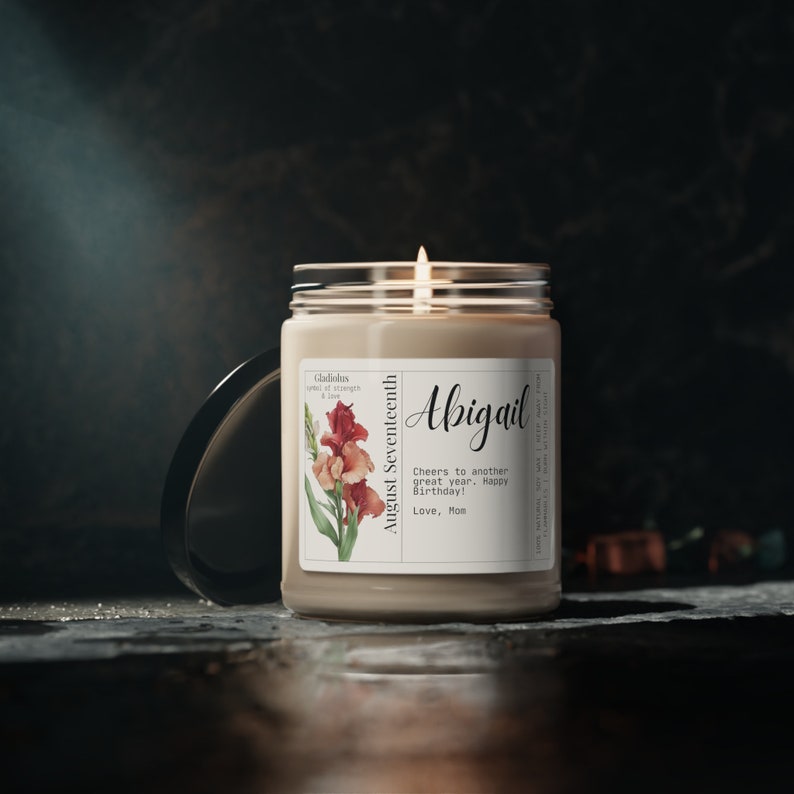 May include: A beige candle with a white label featuring a floral illustration and the text "Gladiolus Symbol of Strength & Love" and "August Seventeenth". The label also includes the name "Abigail" and the message "Cheers to another great year. Happy Birthday! Love, Mom".