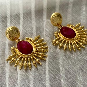 May include: A pair of gold-colored earrings with a sunburst design. Each earring features a textured, round stud and a central, oval-shaped, deep red stone. The sunburst design has small, pointed details radiating outwards.