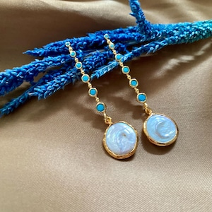 May include: Gold-coloured earrings with a delicate design. Each earring has a chain of small turquoise beads and a larger, round, white pearl-like pendant with a gold border. The earrings are displayed on a neutral background.
