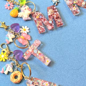 May include: A collection of colorful keychains with gold-colored rings. The keychains feature various charms, including a pineapple, a unicorn, a donut, and letter-shaped pendants filled with glitter and small decorative elements. The background is a solid light blue.