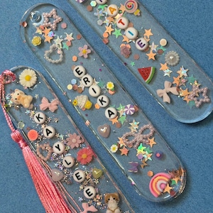 May include: Three clear resin bookmarks with embedded decorative elements. Each bookmark features a name spelled out with letter beads, along with various charms like stars, flowers, and bows. One bookmark has a pink tassel.