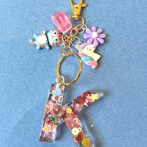 May include: A gold-colored keychain with a letter "K" pendant. The pendant is clear with embedded glitter and floral accents. Additional charms include a blue cat, a pink block, a purple flower, and a unicorn. The keychain has a lobster clasp.