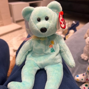 May include: A light blue beanie baby bear with a yellow flower and green leaves embroidered on its chest. The bear has a black nose and black eyes. The bear is wearing a light pink ribbon around its neck. The bear has a red Ty tag attached to its ear with the word "Ty" in white.