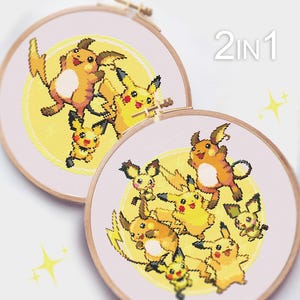 Puede incluir: Dos aros de bordado con patrones de punto de cruz de Pikachu y sus evoluciones. Los aros son de madera y tienen un fondo rosa claro. El texto "2 IN 1" está impreso en negro en el aro superior.