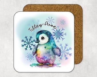 Peut inclure: Un dessous de verre carré avec une illustration à l'aquarelle d'un pingouin coloré, avec le nom "Tilley-Anne" écrit au-dessus. Le pingouin est entouré de flocons de neige bleus sur un fond bleu clair. Le dessous de verre est sur une base en liège marron.