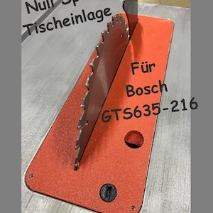 Può includere: Inserzione per sega da tavolo rossa con una lama da sega nera per una sega Bosch GTS635-216. Il testo "Null Spalt Tischeinlage" è scritto sull'inserzione.