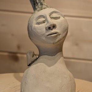 Puede incluir: Un busto de cerámica de color claro que representa una cabeza humana estilizada con los ojos cerrados, con una protuberancia texturizada y curvada hacia arriba en la parte superior de la cabeza y un pequeño elemento triangular en el hombro.