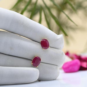 Könnte beinhalten: Ein Paar goldene Ohrstecker, jeweils mit einem großen, oval geschliffenen Rubin-Edelstein. Die Rubine haben eine tiefe, satte rote Farbe und sind in einer einfachen Goldfassung gefasst. Die Ohrringe werden auf einer weißen Oberfläche präsentiert.