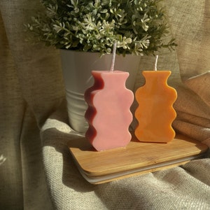May include: Two decorative candles shaped like vases, one pink and one orange, on a wooden base.