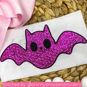 Puede incluir: Un parche de tela blanca con una chauve-souris bordada a máquina con un patrón de textura magenta vibrante similar a un cerebro. El murciélago tiene dos grandes ojos circulares negros sólidos y un contorno cosido en negro. Una cinta rosa es visible en la esquina.