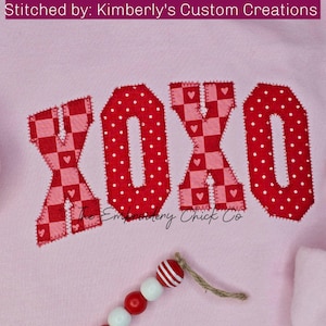May include: A pink sweatshirt with the word "XOXO" stitched on it in red fabric with different patterns. The letters are outlined in red thread.