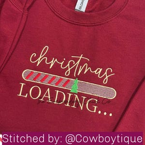 May include: A maroon sweatshirt with the words "christmas LOADING..." embroidered in gold and white thread. The loading bar has red and green stripes with a small green Christmas tree. The text "Stitched by: @Cowboyboutique" is at the bottom.