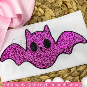 Puede incluir: Un murciélago morado brillante con ojos negros y un diseño cosido. El murciélago está sobre un fondo de tela blanca.