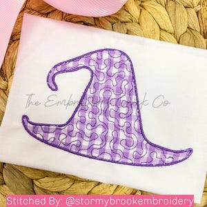 Op de afbeelding: Paarse heksenhoed borduurontwerp op een witte stof. De hoed is een gingham-patroon met een swirl-ontwerp. De tekst "The Embroidery Nook Co." is zichtbaar op de achtergrond.