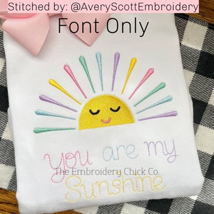 Puede incluir: Diseño bordado en una camisa blanca con un sol amarillo con una cara sonriente y rayos de arcoíris. El texto "You are my Sunshine" está bordado debajo del sol.