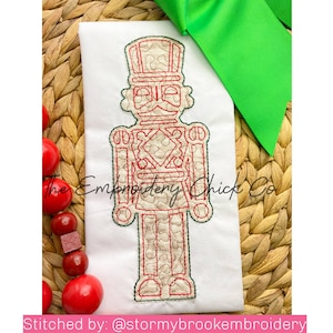 以下が含まれることがあります： 白いTシャツに赤と白の刺繍で描かれたくるみ割り人形のデザイン。デザインは緑色の糸で縁取られています。背景には「The Embroidery Chick Co.」の文字が見えます。