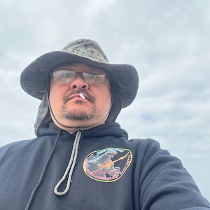 Puede incluir: Un hombre con una sudadera con capucha negra con un gráfico de un monstruo sosteniendo una guitarra. La sudadera tiene el texto "Monster Zero Surf Club". También lleva un sombrero de camuflaje y gafas.