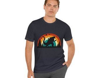May include: A dark blue t-shirt with a retro-style graphic of a monster in front of a sunset. The graphic features a silhouette of a monster with a red, orange, and yellow sunset behind it.