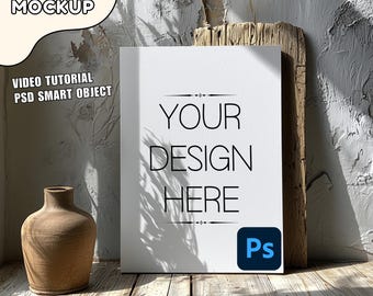 May include: A white canvas mockup with the text "YOUR DESIGN HERE" and a Photoshop logo in the bottom right corner. The canvas is leaning against a wooden plank and a textured wall. A brown clay vase is in the foreground.