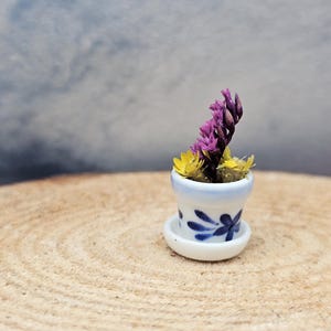 May include: A miniature ceramic flower pot with a blue and white floral design, holding purple and yellow flowers. The pot sits on a matching saucer, placed on a wooden surface.