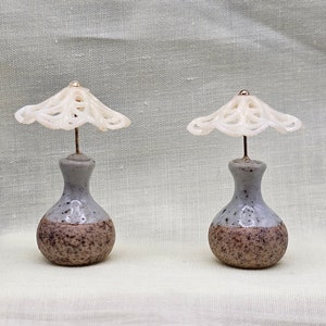 May include: Two miniature ceramic vases with a speckled gray and brown finish, each topped with a delicate, white, lace-like structure. The vases have a small, round base and a slender neck, with a gold-colored bead at the top of the lace structure.