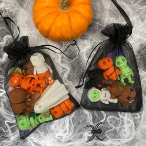 May include: Two black mesh bags filled with Halloween-themed figurines, including pumpkins, skeletons, and ghosts. A small orange pumpkin sits above the bags, and a spider is on the web-like background. The figurines are in various colours.