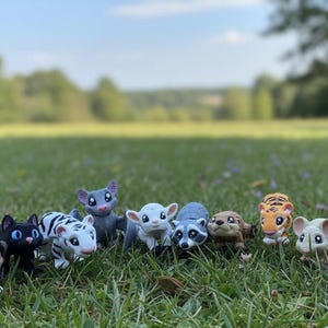May include: A group of animal figurines, such as a dog, cat, and tiger, are displayed on grass. The figurines are brightly coloured and have large eyes, giving them a cartoon-like appearance. The background is blurred, with trees and a blue sky visible.