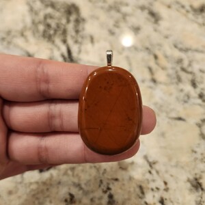 May include: An oval-shaped, polished reddish-brown stone pendant with a smooth, glossy surface and subtle dark brown linear inclusions. The stone is set with a small, silver-toned metal bail at the top for attachment to a necklace chain.