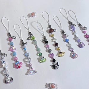 May include: A collection of colorful beaded phone charms with various charms, including stars, hearts, and cartoon characters. The charms feature a variety of pastel colors and clear beads, with silver-tone metal hardware and white cords. Two candy-shaped charms are also present.
