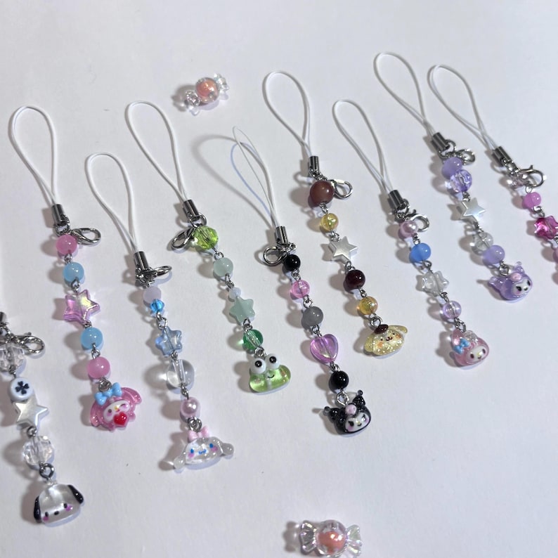 May include: A collection of colorful beaded phone charms with various charms, including stars, hearts, and cartoon characters. The charms feature a variety of pastel colors and clear beads, with silver-tone metal hardware and white cords. Two candy-shaped charms are also present.