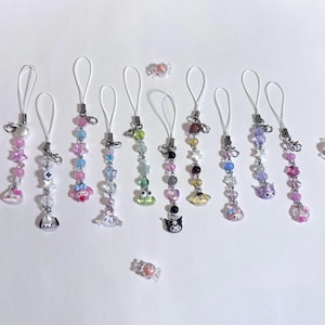 May include: A collection of beaded phone charms in various colors, featuring charms with cartoon characters, stars, and hearts. Each charm has a white loop and silver-colored metal accents. Two small candy-shaped charms are also present.